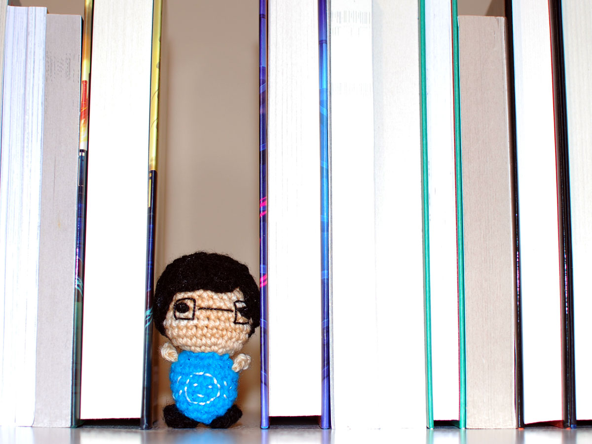 Small crocheted person standing in a gap between several standing books.